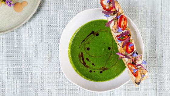 An overhead shot of a bowl of bright green soup with a dark oil drizzle. A long baguette slice sits on the edge, decorated with mussels, red chili rings, and delicate purple and blue floral garnishes. A corner of another plate with a small appetizer is visible in the top left.