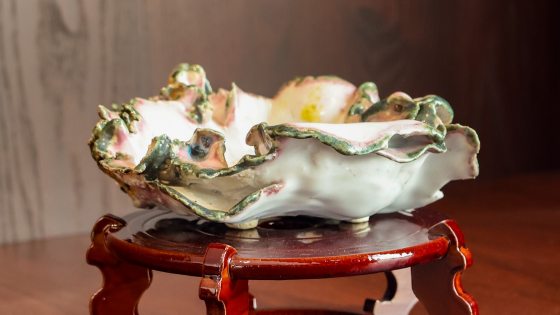 A close-up of an empty, highly ornate Kusamono pot displayed on a dark red, polished wooden stand with curved, decorative legs. The ceramic pot is designed to resemble a delicate, fluted seashell or ruffled flower blossom, featuring a white interior and a heavily textured rim highlighted with soft pink and green glazes.