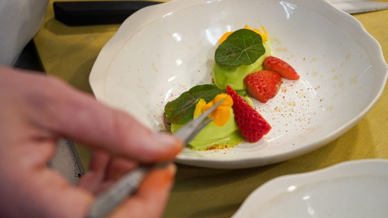 An over-the-shoulder close-up of a chef using kitchen tweezers to place a delicate orange edible flower petal onto a plated dish consisting of vibrant green sorbet, sliced fresh strawberries, and green nasturtium leaves in a wide white ceramic bowl.