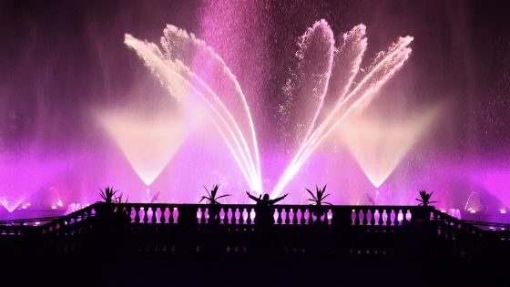 Silhouette of a person standing with arms raised on a balcony, watching a grand, fan-shaped nighttime fountain display illuminated in a deep magenta hue.