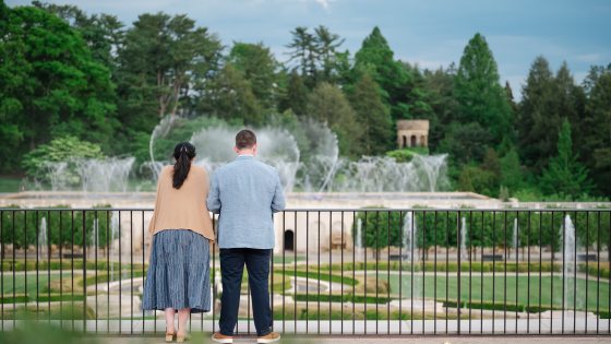 Two people stand at the iron railing of a garden overlook while fountains dance in the background.