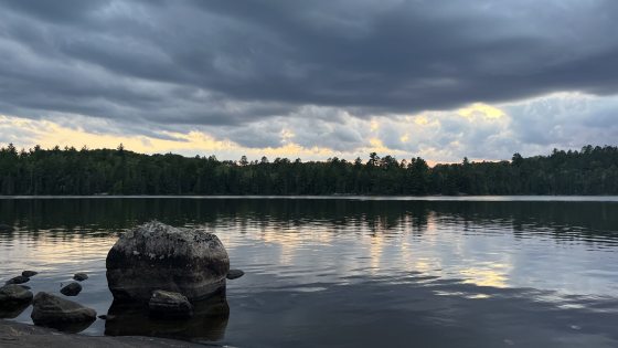 A tranquil lake reflects a dramatic sunset through dark, heavy storm clouds hovering over a distant, densely forested shoreline, with a large rounded boulder resting in the water near the rocky foreground.