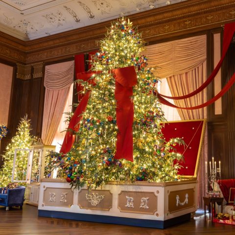 Jewel-toned ornaments shimmer on a heavily decorated 18-foot Fraser fir  that rises from a giant gold and white jewelry box and draped in a sweeping six-foot wide red bow that connects to free standing columns and wraps around the room. 
