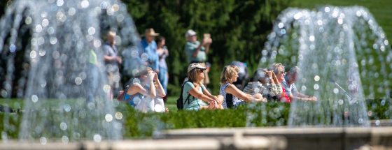 a group of people sitting on a bright green lawn behind a spray if water coming from two fountains