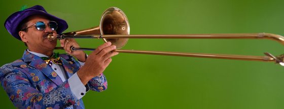 A musician in a colorful floral jacket, sequined bowtie, purple hat, and tinted glasses plays the trombone