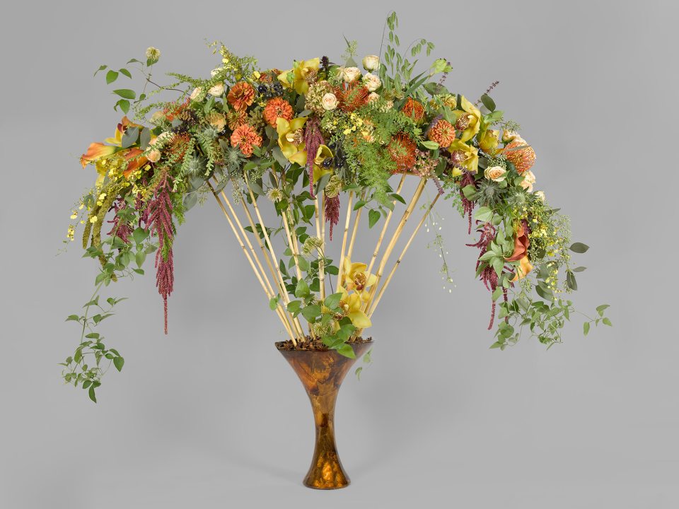 floral arrangement in a brown vase with small bamboo rods and yellow and orange flowers hanging at the top of the rods