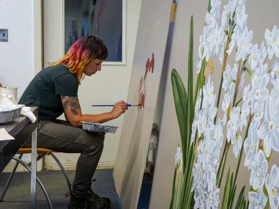 A person in an art studio hunched over painting flowers on a large-scale white board.