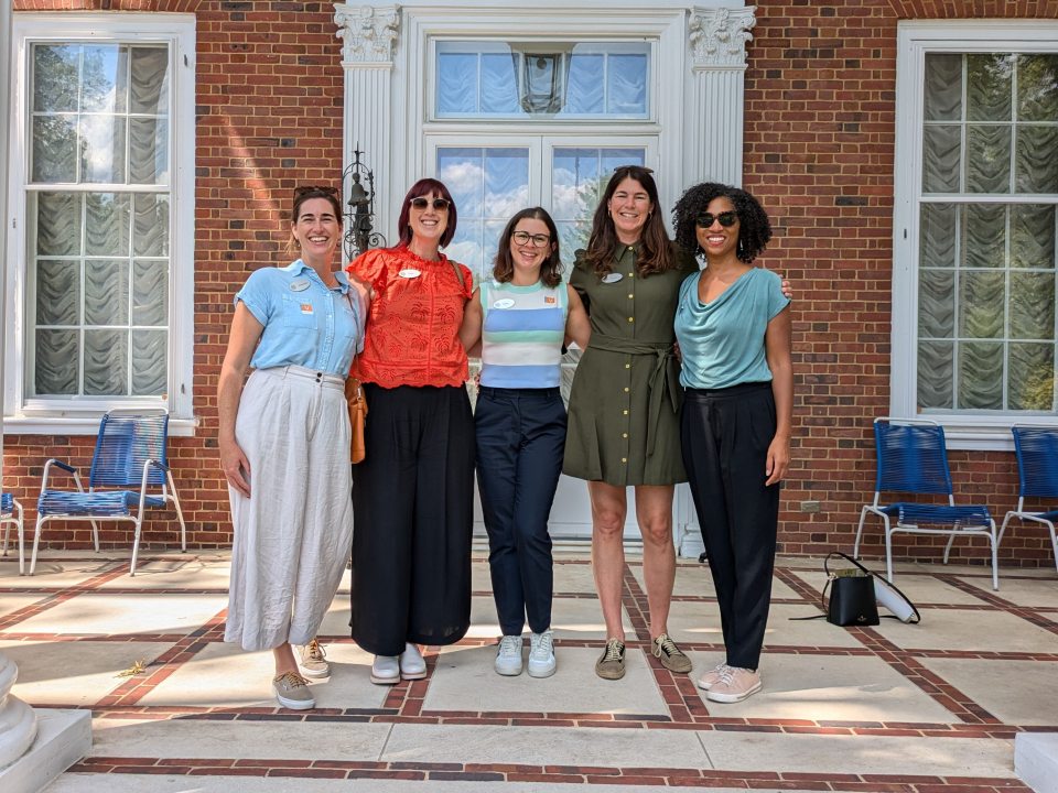 Five people standing shoulder to shoulder in front of a brick building, posing for the camera.