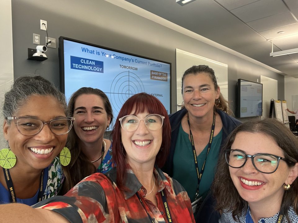A selfie of 5 adults with a classroom in the background. 