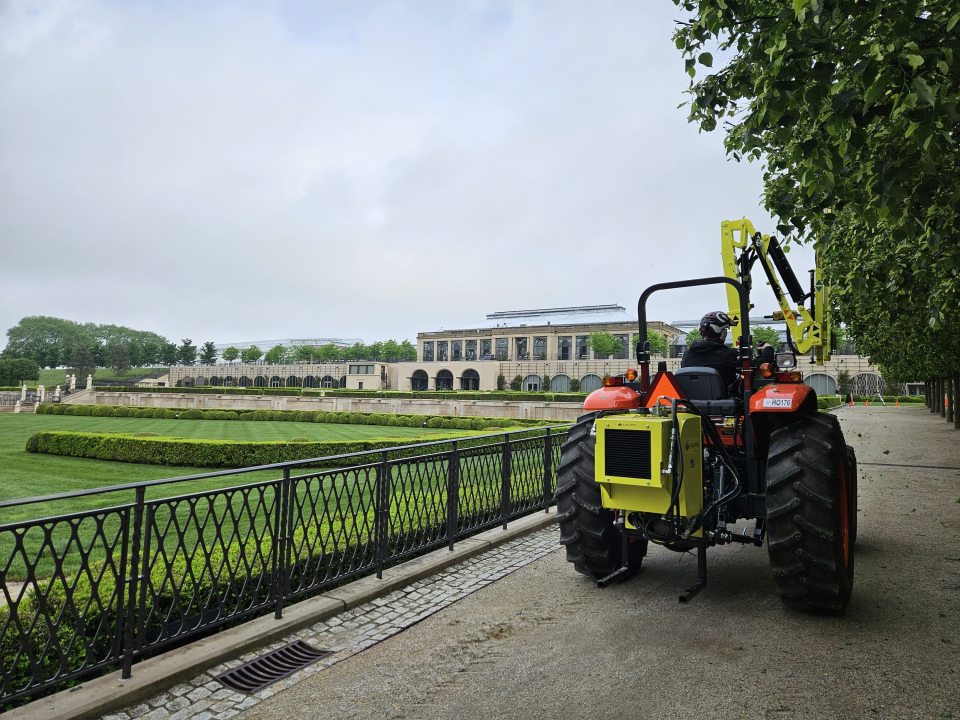 A person in a red tractor trimming trees along an allee with a large yellow shearing device on the front of the tractor.