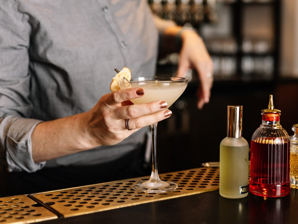 A person's hands behind a bar holding a cocktail glass preparing to hand it to someone. 