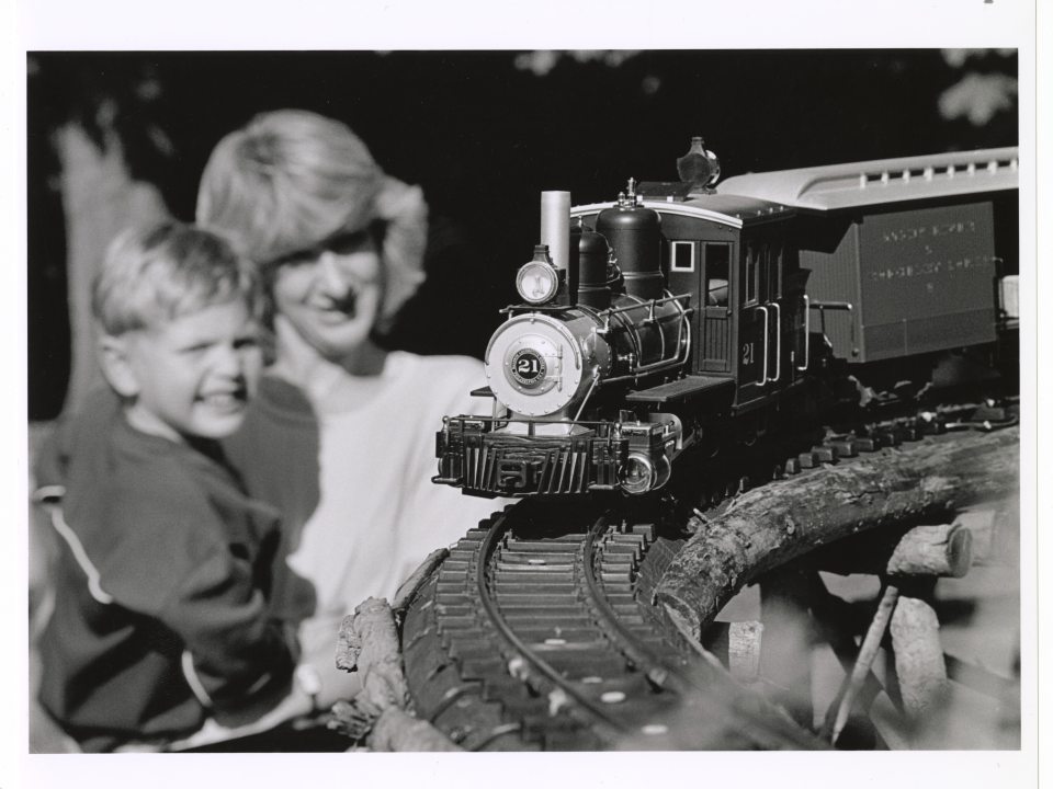 A black and white photograph of a toy or model steam train engine and car running on a curved track in the foreground. In the soft-focus background, an adult woman and a young boy are smiling as they watch the train.