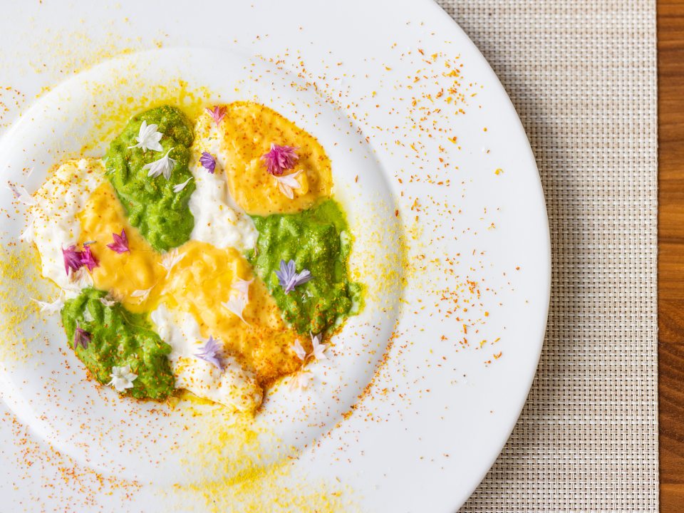 Close-up, overhead view of a plated dish, likely risotto, with alternating dollops of creamy white, vibrant green, and yellow puree or sauce, garnished with edible flowers and surrounded by spice dust.