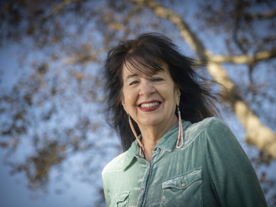 Outdoor portrait of poet Joy Harjo smiling at the camera, wearing a light teal velvet collared shirt and long beaded earrings, with a large tree and clear blue sky out of focus behind her.