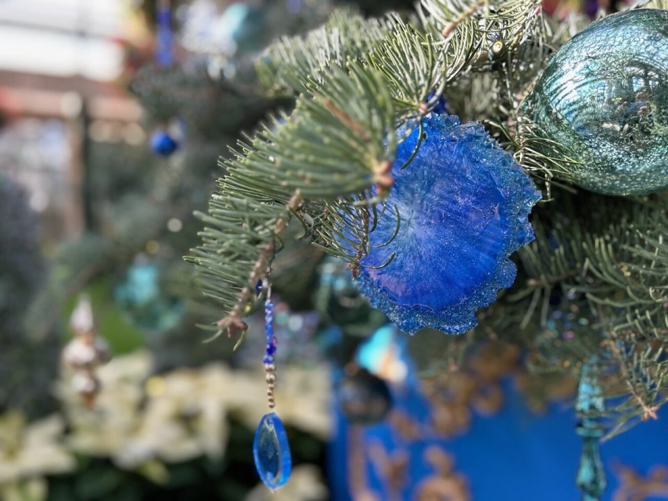 A close-up shot of a Christmas tree branch adorned with blue and turquoise ornaments. A striking, fuzzy, royal blue floral or star-shaped ornament is visible, along with a hanging decoration featuring a dangling, tear-drop-shaped clear blue crystal bead. White poinsettias are visible and slightly blurred in the lower left background