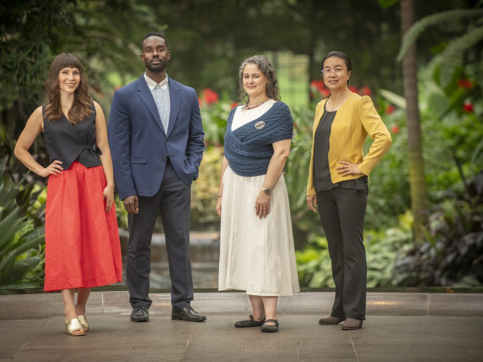 A full-body group portrait of four Longwood Fellows standing side-by-side in formal business-casual attire against a lush green background.