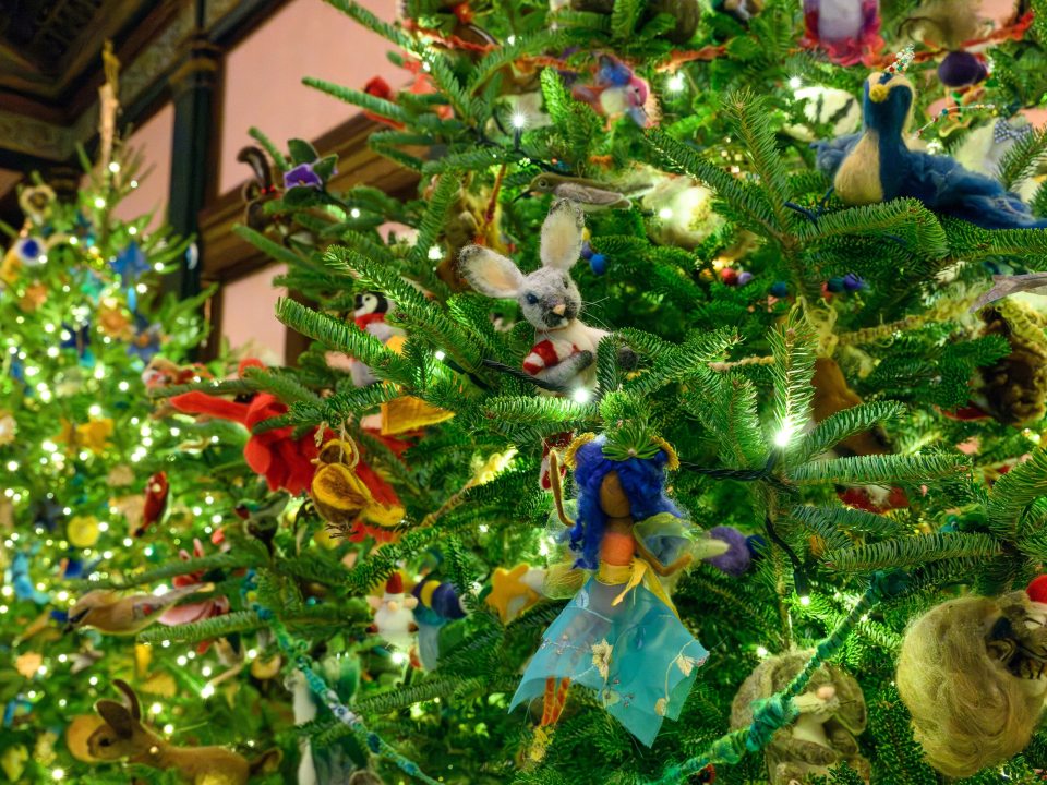 A vertical shot of a heavily decorated Christmas tree. The ornaments are unique, including a central needle-felted white rabbit with a red scarf, a figure with a blue fabric skirt, a felted blue bird, and various other whimsical animal ornaments.