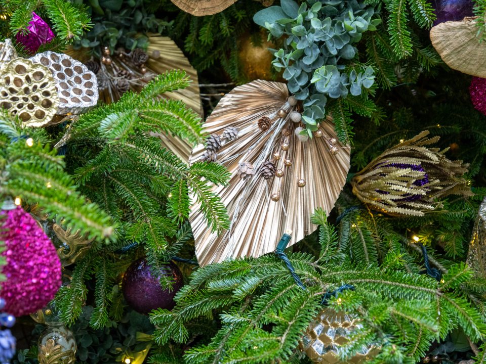 A close-up of a richly decorated Christmas tree showing a variety of ornaments, including large, pleated, metallic gold fan-shaped decorations, natural elements like pine sprigs and small pinecones, and bright, glittery ornaments in shades of magenta, purple, and gold.