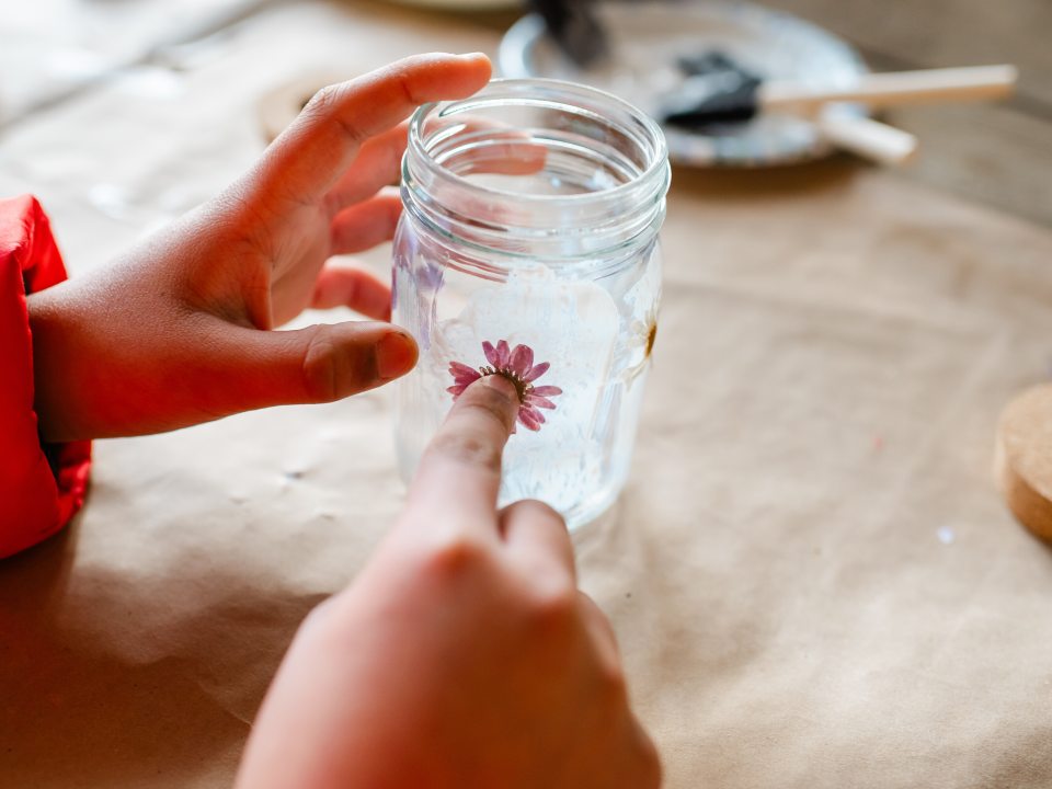 A close-up shot of hands carefully pressing a small, pink dried flower onto the side of a glass jar, showing the intricate detail of the craft project.