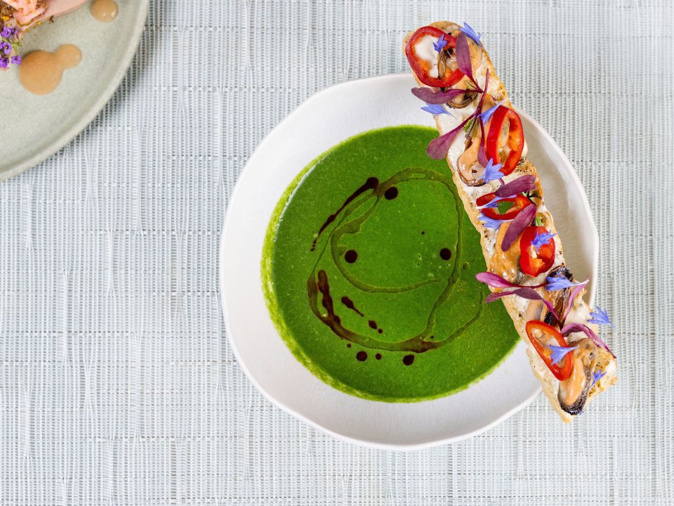 An overhead shot of a bowl of bright green soup with a dark oil drizzle. A long baguette slice sits on the edge, decorated with mussels, red chili rings, and delicate purple and blue floral garnishes. A corner of another plate with a small appetizer is visible in the top left.