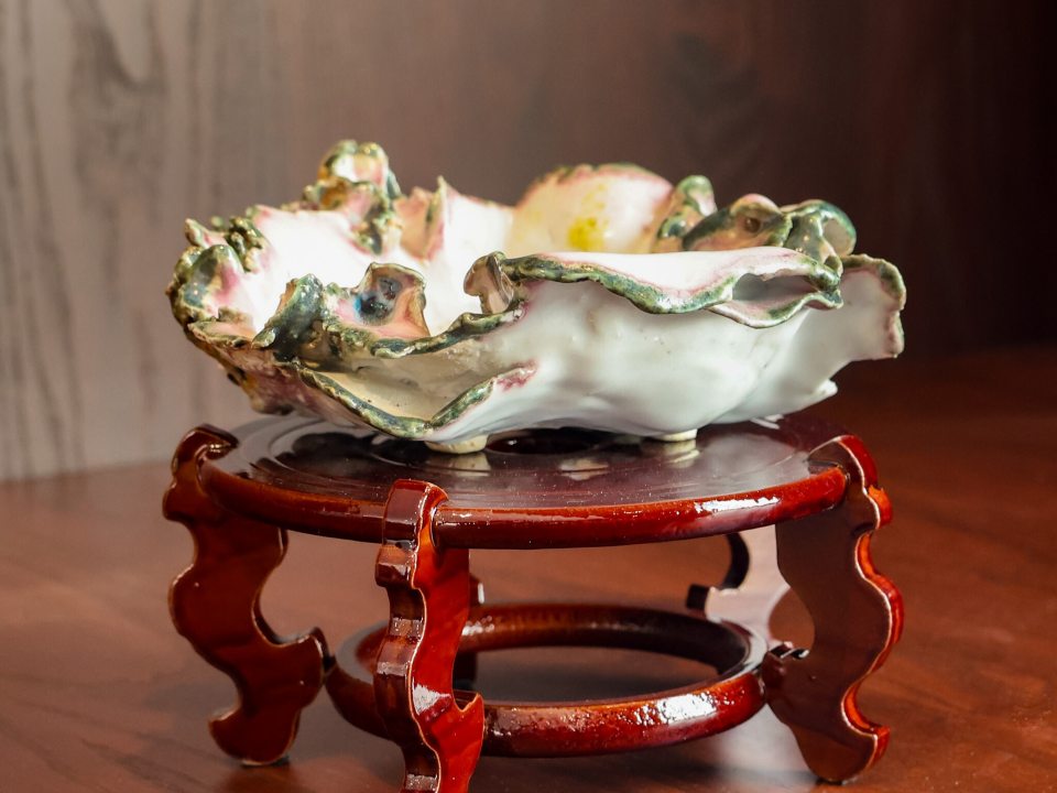A close-up of an empty, highly ornate Kusamono pot displayed on a dark red, polished wooden stand with curved, decorative legs. The ceramic pot is designed to resemble a delicate, fluted seashell or ruffled flower blossom, featuring a white interior and a heavily textured rim highlighted with soft pink and green glazes.
