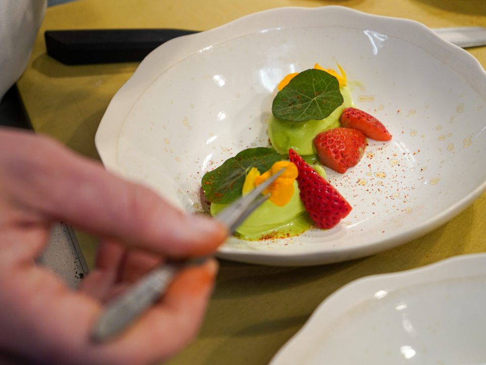 An over-the-shoulder close-up of a chef using kitchen tweezers to place a delicate orange edible flower petal onto a plated dish consisting of vibrant green sorbet, sliced fresh strawberries, and green nasturtium leaves in a wide white ceramic bowl.