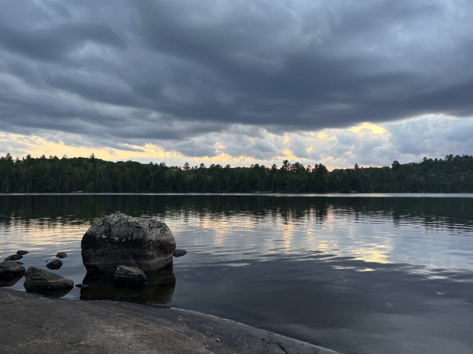 A tranquil lake reflects a dramatic sunset through dark, heavy storm clouds hovering over a distant, densely forested shoreline, with a large rounded boulder resting in the water near the rocky foreground.