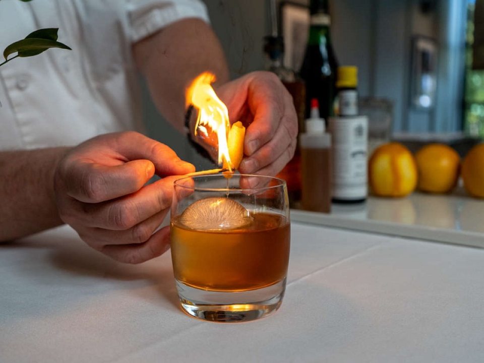 a glass of alcohol with a set of hands lighting a lemon peel on fire 