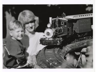 A black and white photograph of a toy or model steam train engine and car running on a curved track in the foreground. In the soft-focus background, an adult woman and a young boy are smiling as they watch the train.