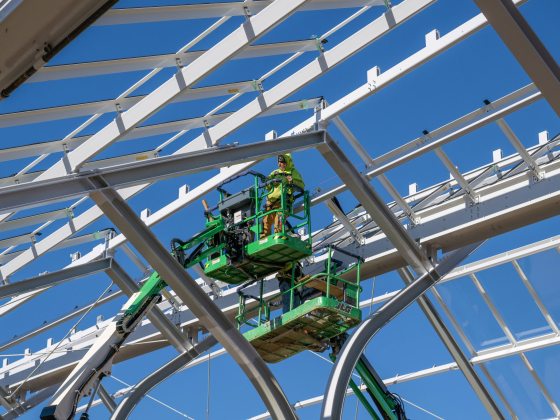 a construction worker atop a large crane is framed by steel arches against a backdrop of blue sky