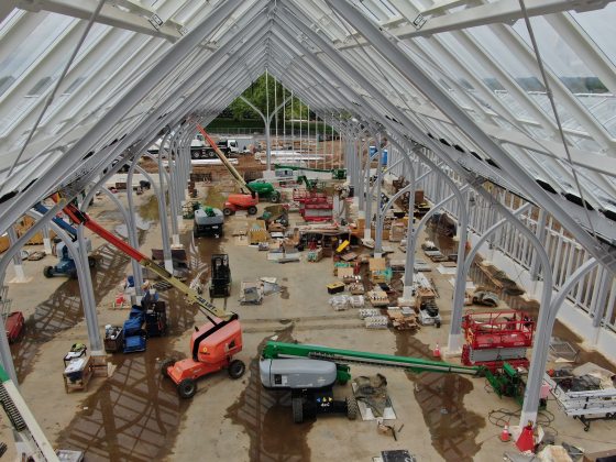 Steel beams and glass of a greenhouse under construction frame a multitude of construction equipment beneath