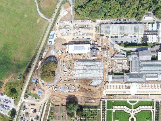 Overhead view of West Conservatory construction project, showing Main Fountain Garden in right foreground and existing Main Conservatory directly behind it, with greenhouse construction zone to its left, at center of photograph.
