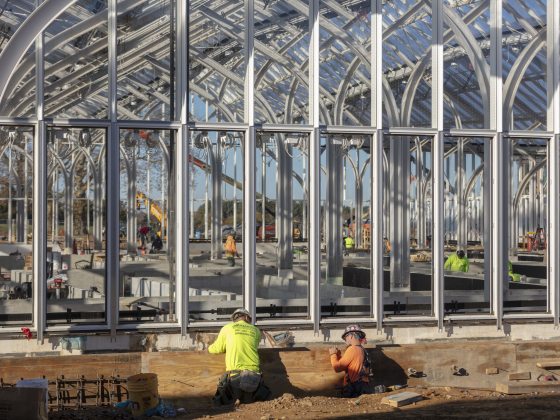 Construction workers amid an open framework of arched metal columns and beams.