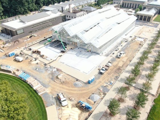 A large conservatory under construction, with a long ceiling of four rows of arched glass, new plantings of trees to the outside right, an empty waterlily courtyard and older conservatory in the background, and a large office building, boiler room, and production greenhouses in a long row of glass triangles at the left of the image.