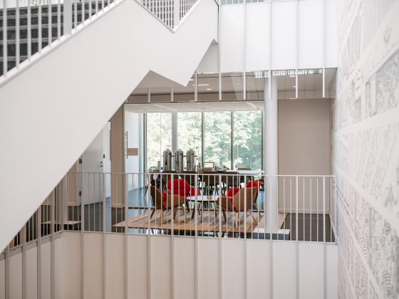 Interior image of white angular stairway and white vertical bars framing a room with a table and red chairs.