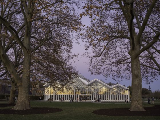 Exterior view of angular glass house against a purple sky at dusk, with large trees and a lawn in the foreground.