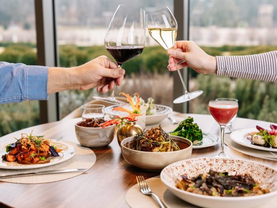 A table is set with a variety of beautifully presented food dishes, with one hand reaching across from each side of the table, clinking glasses of red and white wine.