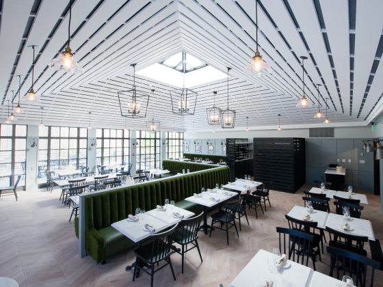 Diagonal view of brightly lit dining space with beamed ceiling and dark green plush benches, white tables, and dark wood chairs.