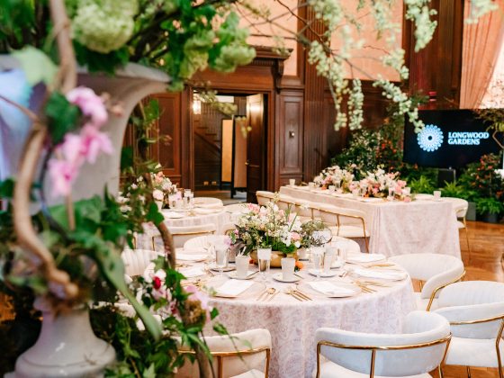 Elegant dining setup at Longwood Gardens, featuring beautifully arranged tables with floral centerpieces, in a richly decorated room with visible Longwood Gardens branding in the background