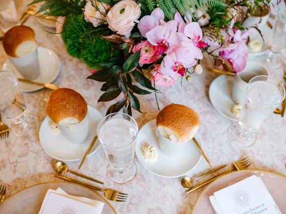 Elegant dining table setting featuring plates with rolls, glasses of water, golden cutlery, and a center arrangement of pink orchids and greenery. Printed menus and napkins are neatly placed on each plate.