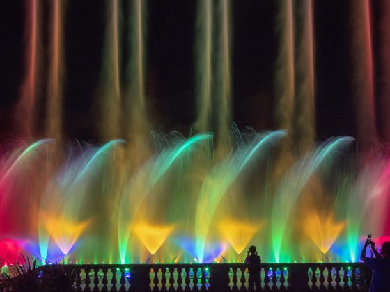 A row of parallel fountain pairs in rainbow colors shoot toward the sky, with a middle layer of dancing fountain spray, and a lower level of cone-shaped fountains.