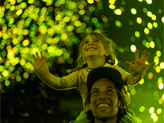 A joyfully smiling child rides on the shoulders of a smiling adult as they gaze upward at yellow-green sparks of light.