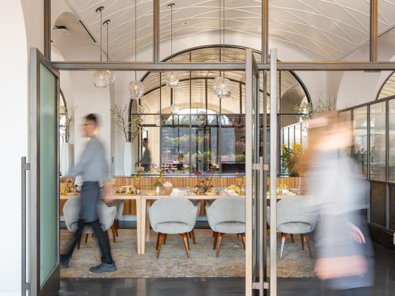 View through open doors into a dining area, set for service, against a backdrop of arched windows, with a server walking to the left.