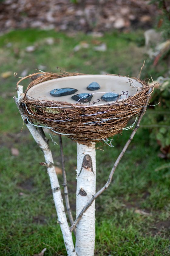 blues stones covered by water in a natural bird bath contained in a nest of woven brown twigs and wire, elevated on a pole of white birch
