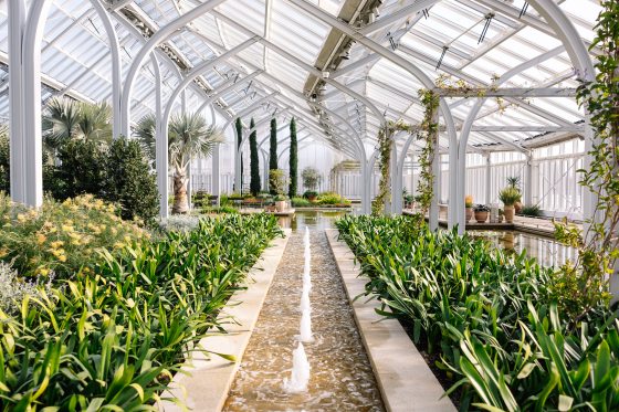 The indoor of a conservatory with a fontain running through the center and plants in beds on either side.