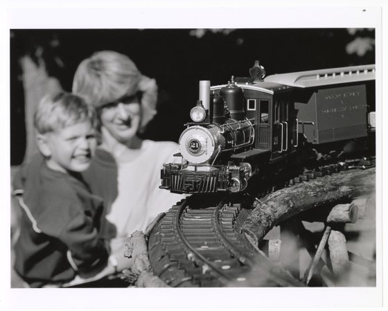 A black and white photograph of a toy or model steam train engine and car running on a curved track in the foreground. In the soft-focus background, an adult woman and a young boy are smiling as they watch the train.
