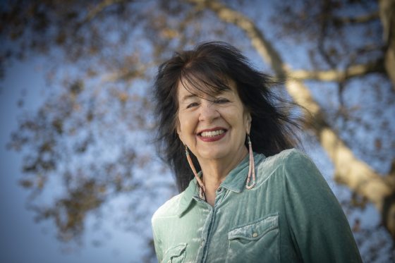 Outdoor portrait of poet Joy Harjo smiling at the camera, wearing a light teal velvet collared shirt and long beaded earrings, with a large tree and clear blue sky out of focus behind her.