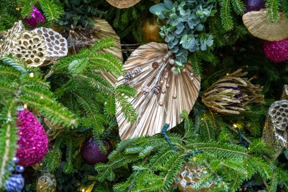 A close-up of a richly decorated Christmas tree showing a variety of ornaments, including large, pleated, metallic gold fan-shaped decorations, natural elements like pine sprigs and small pinecones, and bright, glittery ornaments in shades of magenta, purple, and gold.