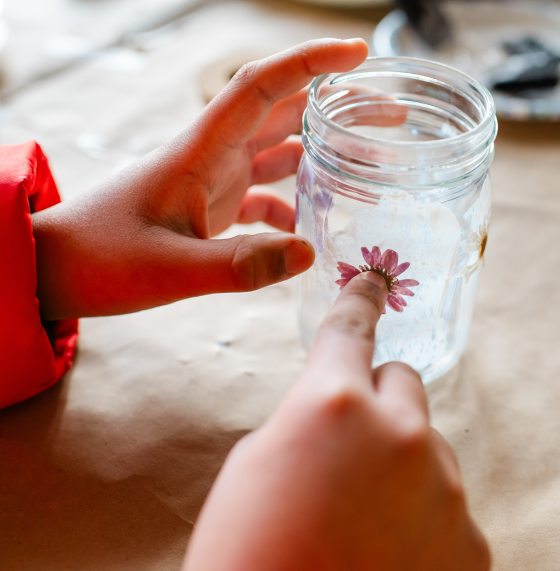 A close-up shot of hands carefully pressing a small, pink dried flower onto the side of a glass jar, showing the intricate detail of the craft project.