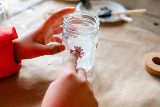 A close-up shot of hands carefully pressing a small, pink dried flower onto the side of a glass jar, showing the intricate detail of the craft project.